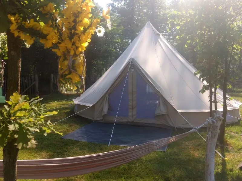 Lichtgekleurde bell tent op een grasveld tussen de bomen, met een hangmat en een pot met gele bloemen op de voorgrond. Het zonlicht valt sfeervol door de bladeren.