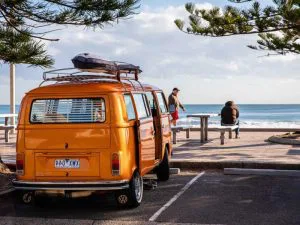 Oranje camper geparkeerd aan de kust met uitzicht op zee, terwijl twee mensen op een bankje naar de golven kijken.