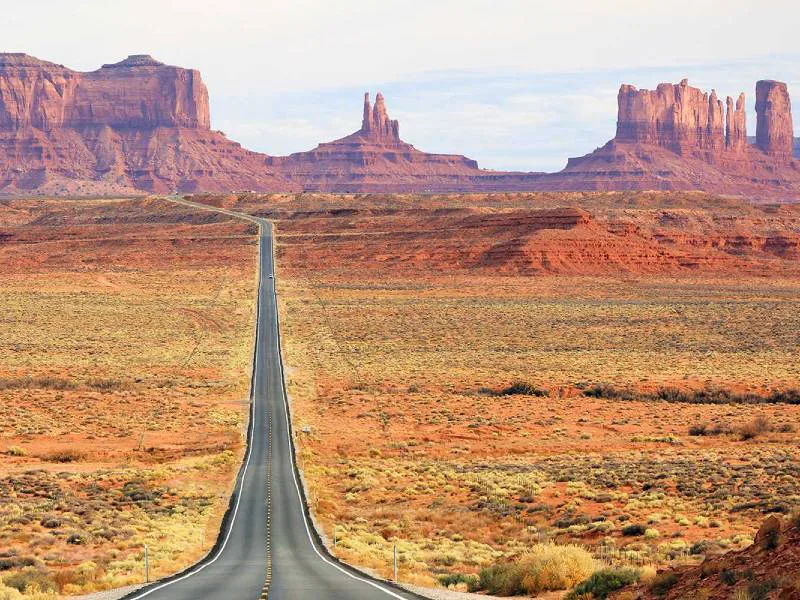 Rechte weg door een droog woestijnlandschap met roodbruine rotsformaties en plateaus in de verte.