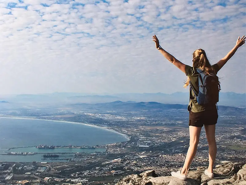 Vrouw met rugzak staat op een bergtop met haar armen in de lucht en kijkt uit over de kustlijn, de zee en de stad beneden.