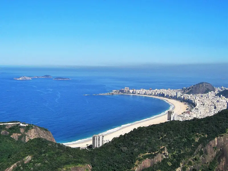Uitzicht vanaf een groene heuvel op de kustlijn van Rio de Janeiro, met de helderblauwe zee, het uitgestrekte strand van Copacabana en de stad op de achtergrond.