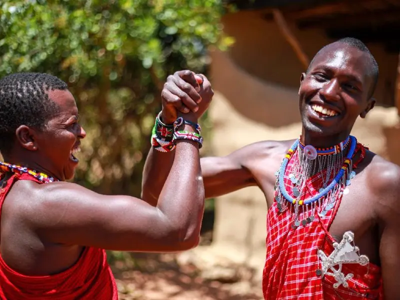 Twee mannen in traditionele Maasai-kleding lachen en schudden elkaars hand in een vrolijk gebaar.