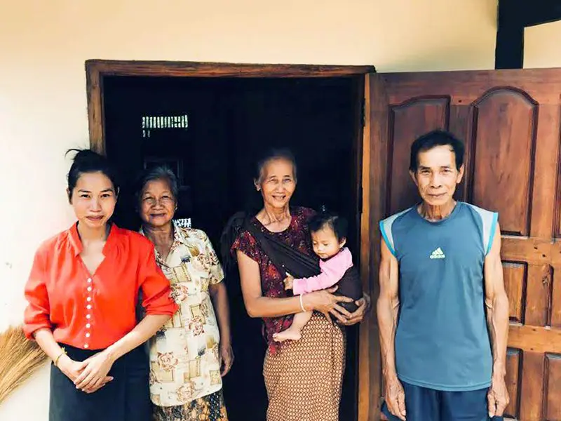 Vier volwassenen en een baby staan lachend voor de ingang van een huis, met een open houten deur naast hen.