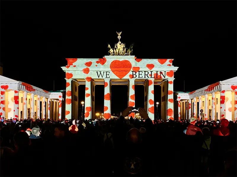 De Brandenburger Tor in Berlijn, ’s avonds verlicht met rode hartjes en de tekst "WE ♥ BERLIN", terwijl een grote menigte toeschouwers toekijkt.