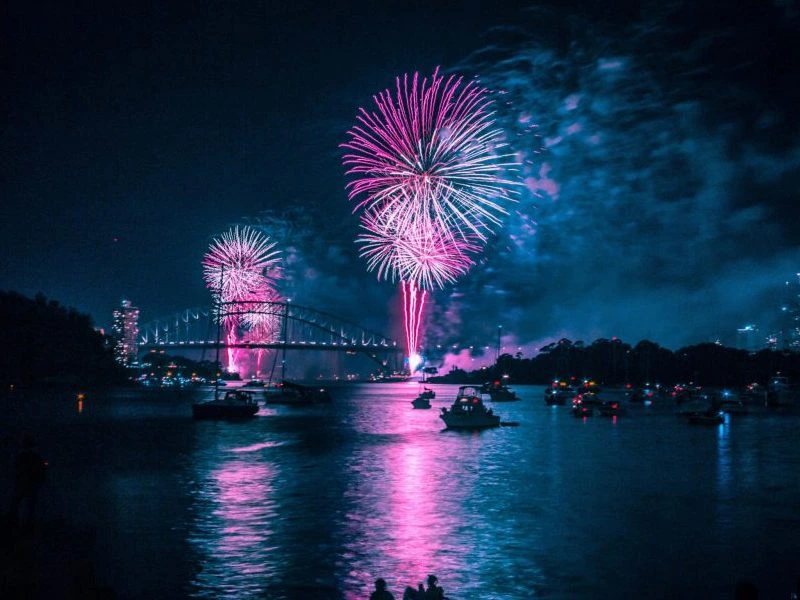 Roze en paarse vuurpijlen verlichten de nachtelijke lucht boven een brug, met boten op het water die de reflecties van het vuurwerk weerkaatsen.