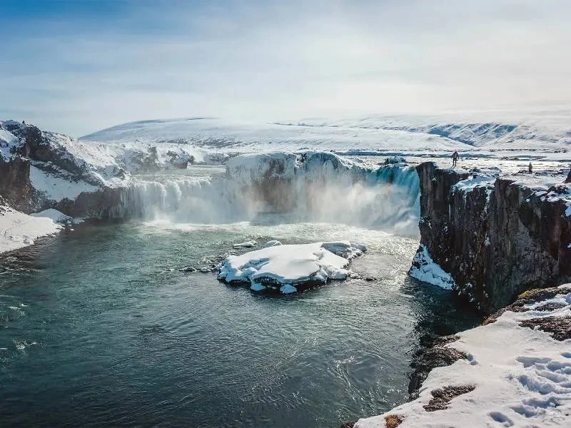 Bevroren waterval in IJsland, omgeven door besneeuwde rotsen en heuvels, met opspattend water en enkele mensen die op de klifrand staan.