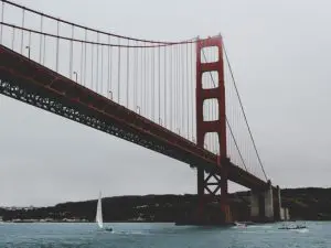 Rode hangbrug die over een brede rivier loopt met zeilboten op het water en heuvels op de achtergrond onder een grijze lucht.
