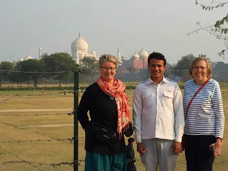 Twee vrouwen en een man poseren lachend voor een hek met prikkeldraad, met de Taj Mahal zichtbaar op de achtergrond.