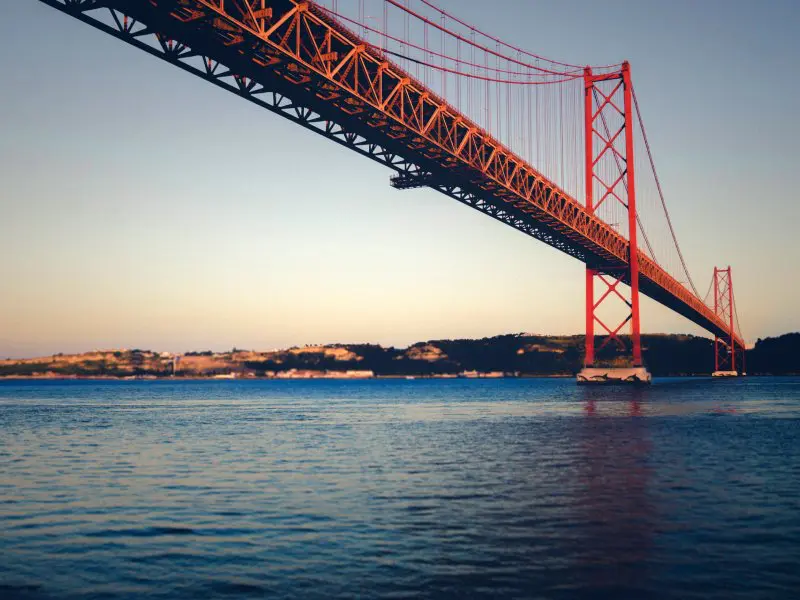 Rode hangbrug over een brede rivier met heuvelachtig landschap op de achtergrond bij zonsondergang.