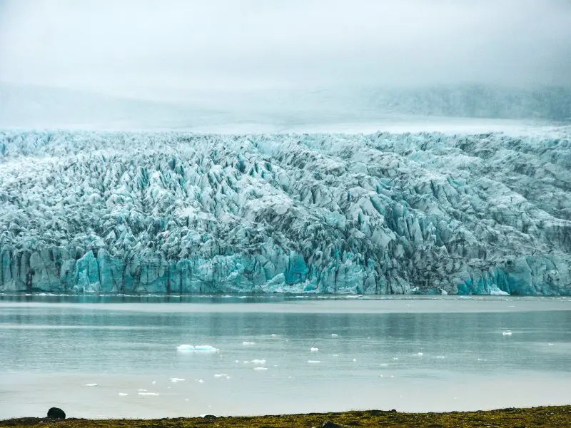 Uitzicht op een massieve blauwe gletsjer die uitkomt in een rustige lagune met drijvende ijsbrokken.
