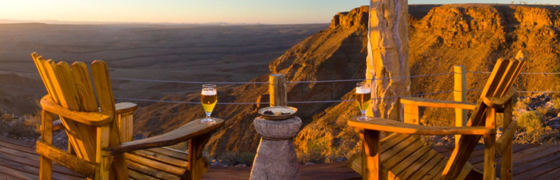 Twee houten stoelen met glazen bier op een terras, uitkijkend over een ruig berglandschap in het warme licht van de ondergaande zon.