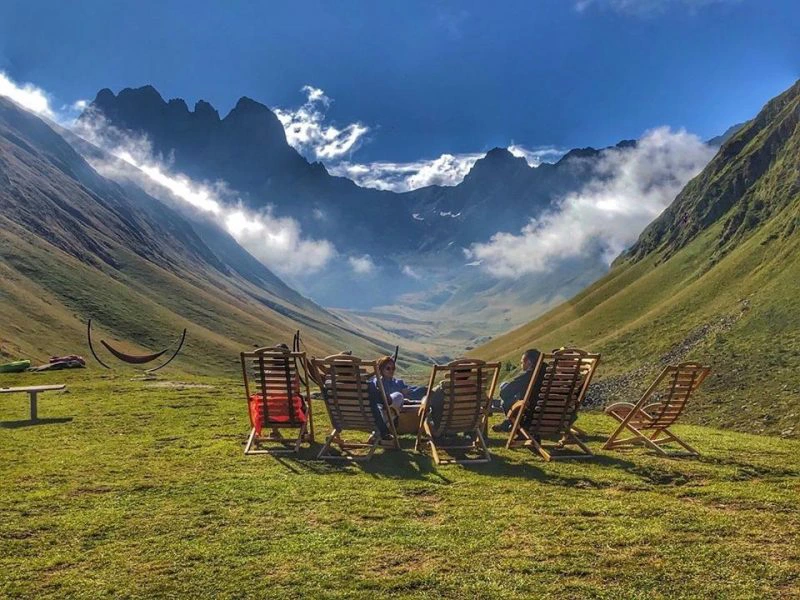 Mensen zitten in ligstoelen op een grasveld, uitkijkend over een groene vallei met hoge bergen en wolken op de achtergrond.