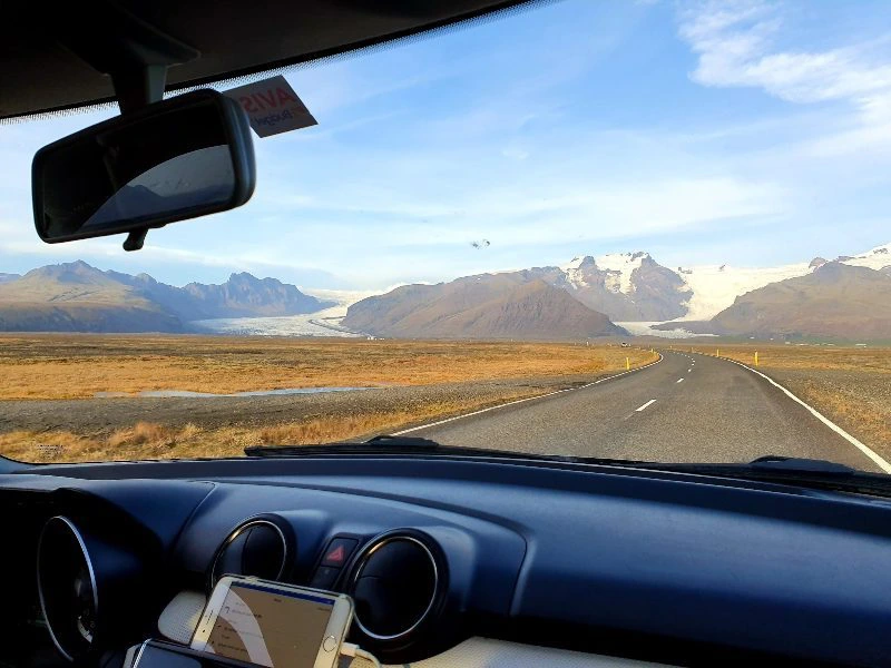 Uitzicht vanuit een rijdende auto op een lege weg die door een weids IJslands landschap slingert, met op de achtergrond besneeuwde bergen en een gletsjer.