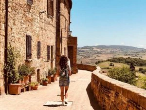 Vrouw loopt met een rugzak door een zonovergoten straatje langs een oud stenen gebouw, met uitzicht op het Toscaanse heuvellandschap.