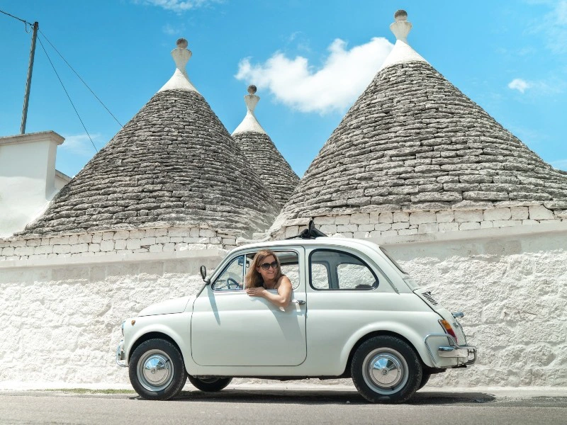 Vrouw in een witte oldtimer rijdt langs traditionele trulli-huizen met kegelvormige daken in Puglia, Italië.