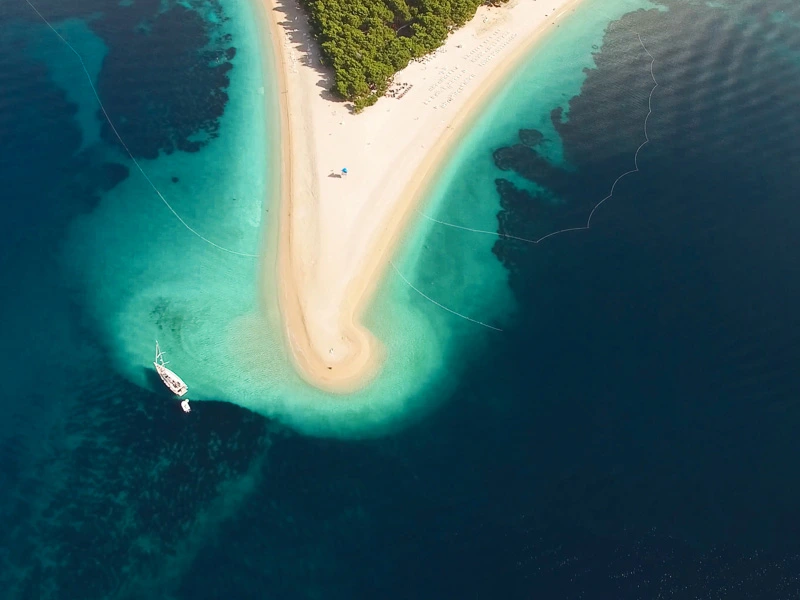 Luchtfoto van een smalle zandstrook die uitloopt in zee, omringd door turquoise water met een zeilboot dichtbij de kust.