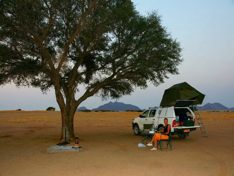 Kampeerplek in de woestijn met een 4x4 auto met daktent onder een boom, een persoon zit ontspannen op een campingstoel met bergen op de achtergrond.