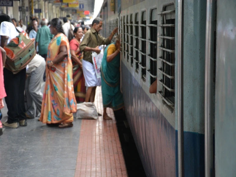 Mensen in kleurrijke kleding op een druk treinstation in India, waar een vrouw door het raam van een blauwe trein naar binnen reikt.