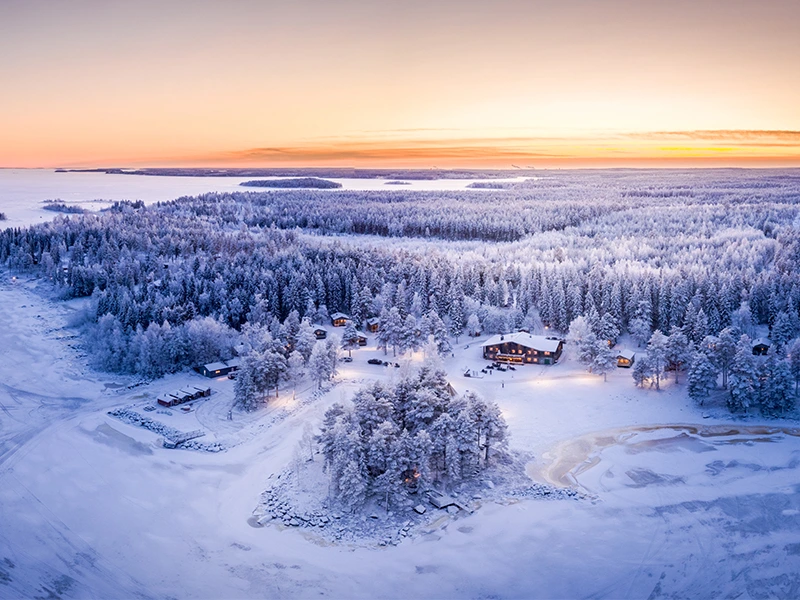 Luchtfoto van een winters landschap in Lapland met besneeuwde bossen, houten huisjes en een oranje-roze gloed van de ondergaande zon aan de horizon.