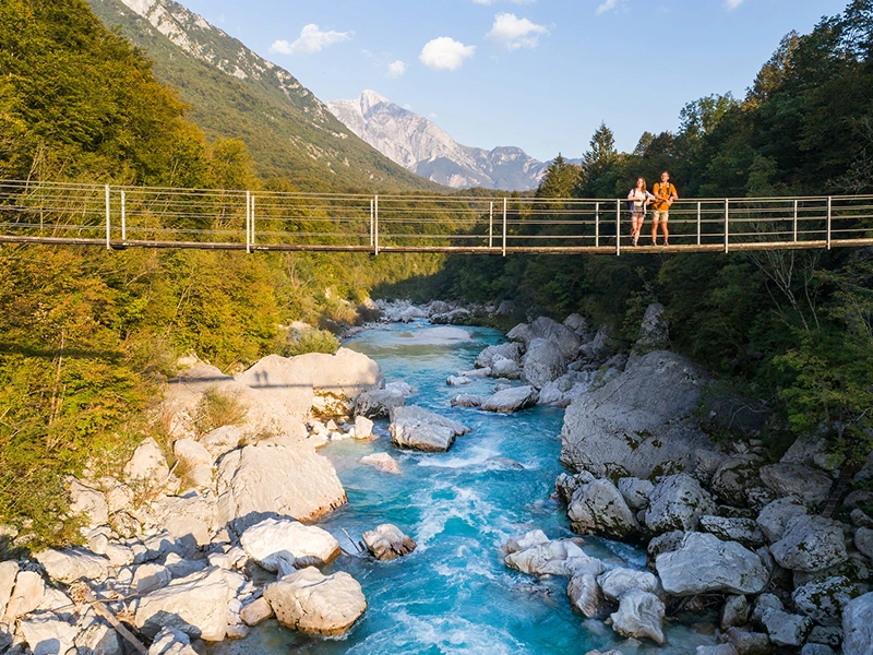 Twee mensen staan op een hangbrug boven een helderblauwe rivier, omringd door groene bossen en bergen.