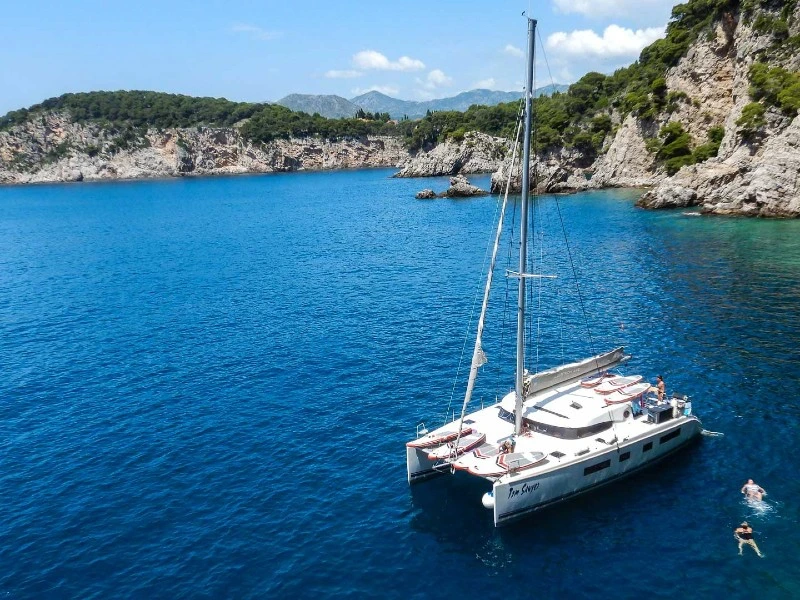 Witte catamaran ligt voor anker in een blauwe baai, omringd door groene rotsen, terwijl mensen in het water zwemmen.