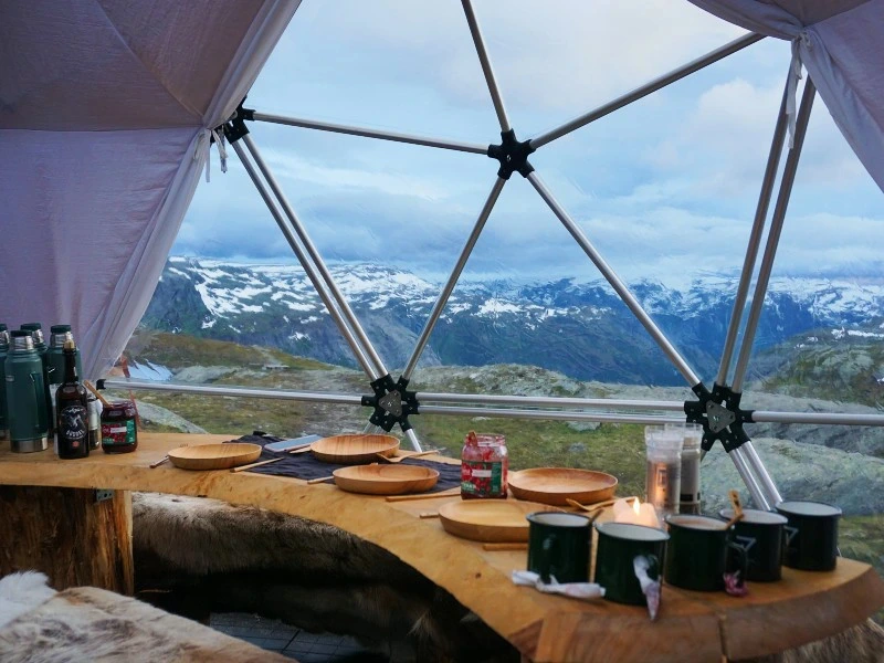 Houten tafel met borden, mokken en thermoskannen in een glamping koepeltent met uitzicht op besneeuwde bergen.