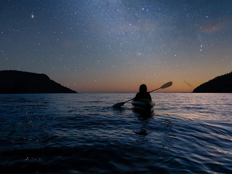Een persoon in een kajak peddelt over kalm water bij zonsondergang, met een sterrenhemel vol twinkelende sterren boven zich.