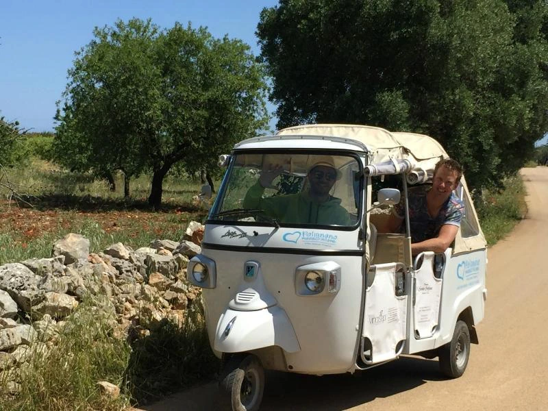 Man leunt uit een witte tuk-tuk op een landweggetje, met een bestuurder binnenin en groene bomen op de achtergrond.