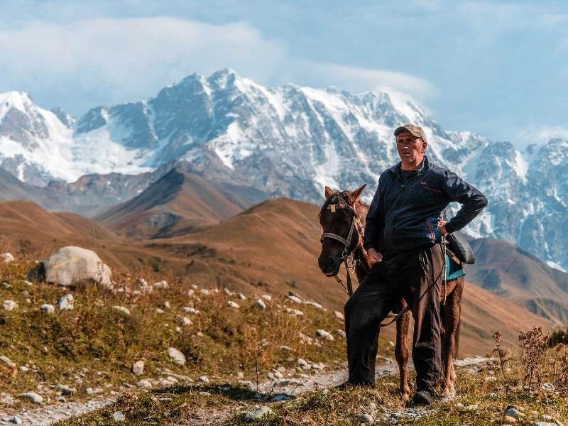 Man met een paard staat in een berglandschap met besneeuwde toppen op de achtergrond.