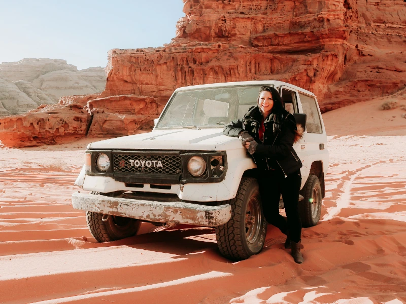 Vrouw leunt lachend tegen een witte Toyota jeep in een rood woestijnlandschap met indrukwekkende rotsformaties.