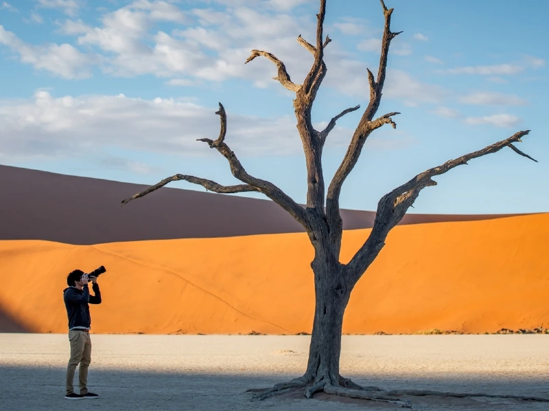 Fotograaf maakt een foto van een dorre, kale boom in de witte kleivlakte van Deadvlei, met hoge oranje zandduinen op de achtergrond in Namibië.