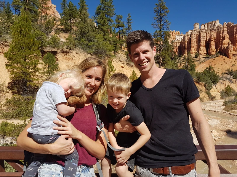 Gezin met twee jonge kinderen poseert lachend voor roodkleurige rotsformaties en dennenbomen in een nationaal park.