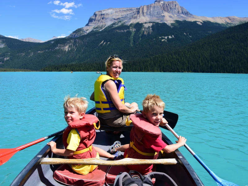 Twee jonge jongens en een vrouw kanoën op een turquoise meer, met hoge bergen en een dicht bos op de achtergrond onder een blauwe lucht.