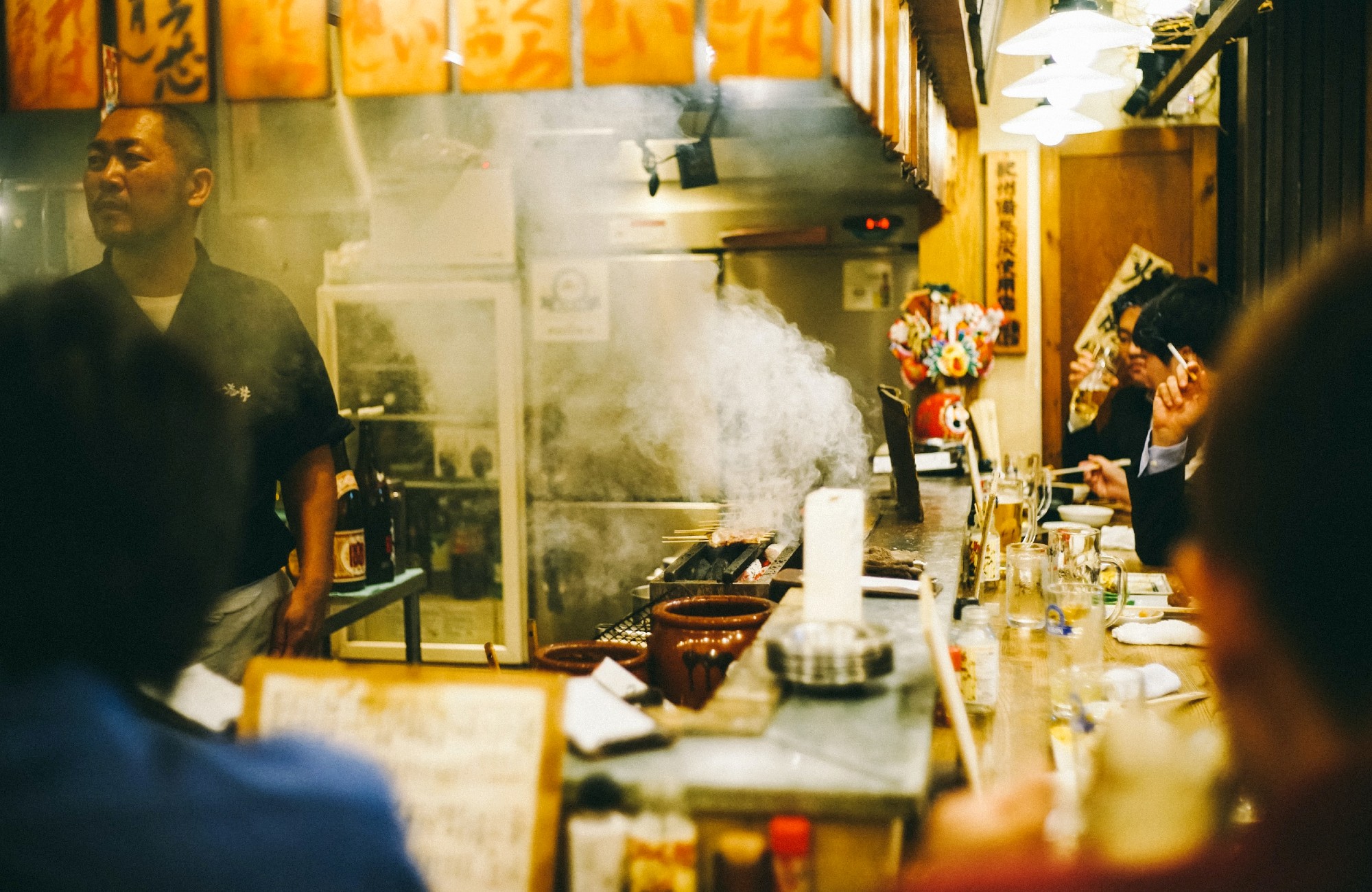 Kleine Japanse eetbar met sissende grill, rokende gerechten en gasten die aan de bar eten en drinken.