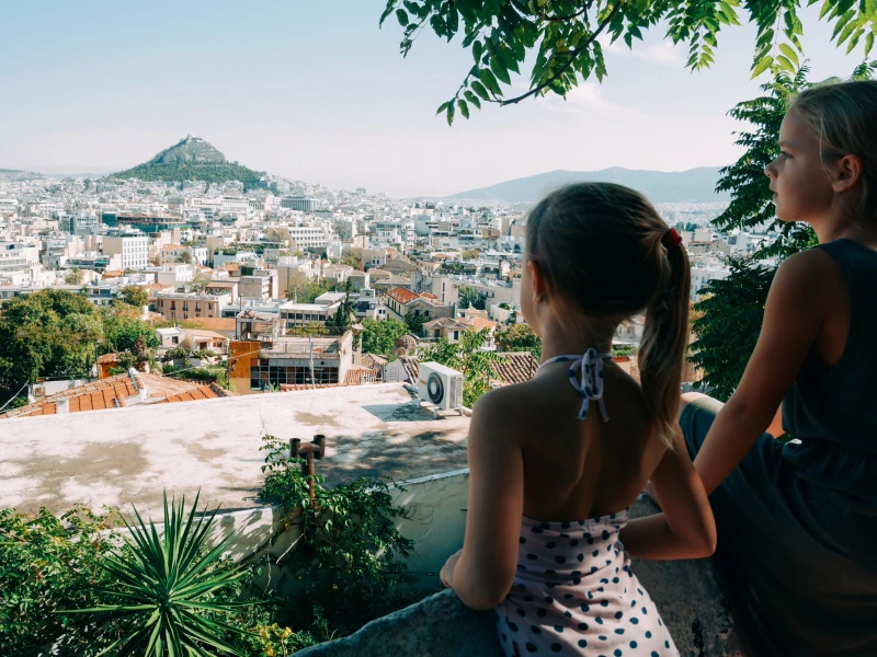 Twee meisjes kijken uit over de stad Athene met de groene heuvel van Lycabettus op de achtergrond, omringd door zonovergoten daken en mediterrane begroeiing.