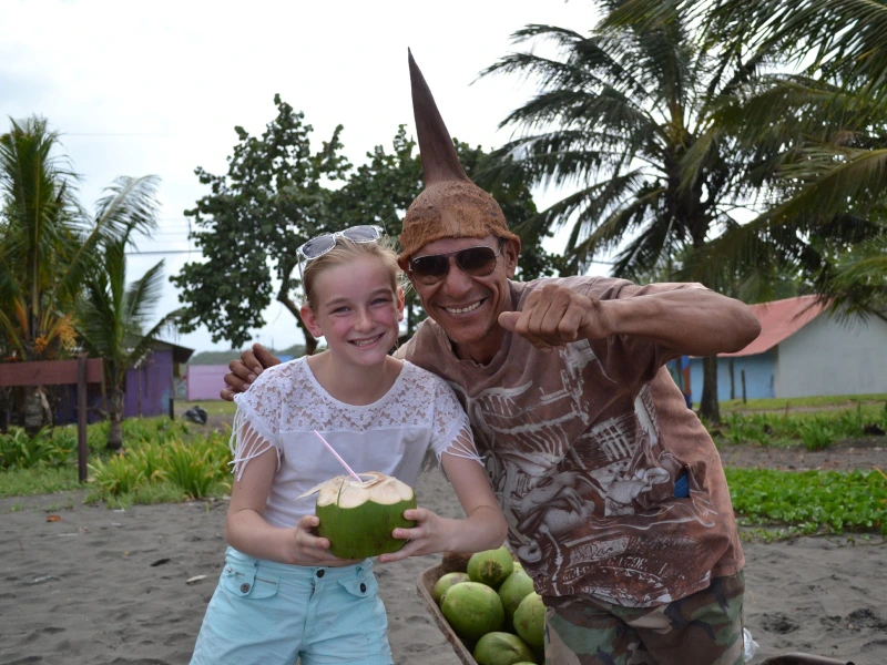 Meisje met een kokosnoot met rietje en een lachende man met zonnebril en kokoshoed, samen poserend op een strand met palmbomen op de achtergrond.