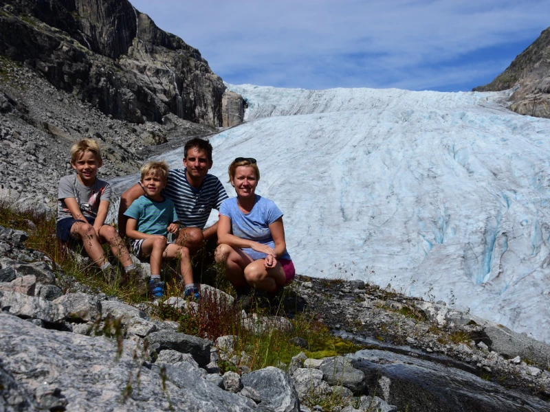 Gezin van vier zit op rotsen voor een indrukwekkende gletsjer, omringd door bergen en een strakblauwe lucht.