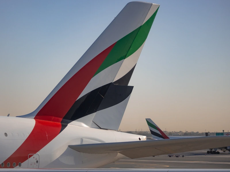 Close-up van het staartstuk van een Emirates-vliegtuig met de kenmerkende rood-groen-zwart-witte kleuren, geparkeerd op het vliegveld bij zonsopkomst of -ondergang.