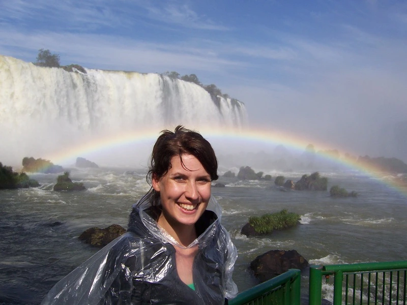 Vrouw in een regenponcho staat lachend voor de Iguazu-watervallen, met een regenboog in de nevel achter haar.