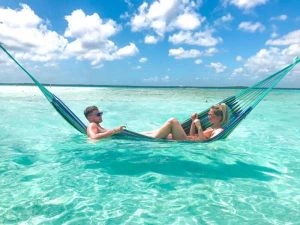 Twee mensen liggen samen in een hangmat boven helder turquoise zeewater met een blauwe lucht en witte wolken erboven