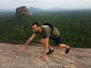 Man klimt een rots op met uitzicht over een uitgestrekt groen boslandschap en de rotsformatie Sigiriya in Sri Lanka op de achtergrond.