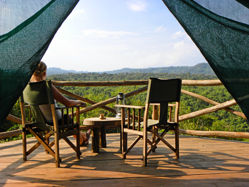 Vrouw zit in een safaristoel op een houten veranda met uitzicht op een groene jungle, naast haar een lege stoel en een tafeltje met een lamp en een mok.