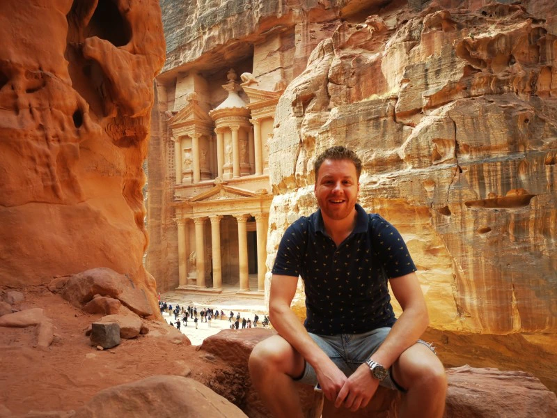 Lachende man zit op een rots met uitzicht op de beroemde Schatkamer van Petra, een uit rotsen uitgehouwen tempel in Jordanië.