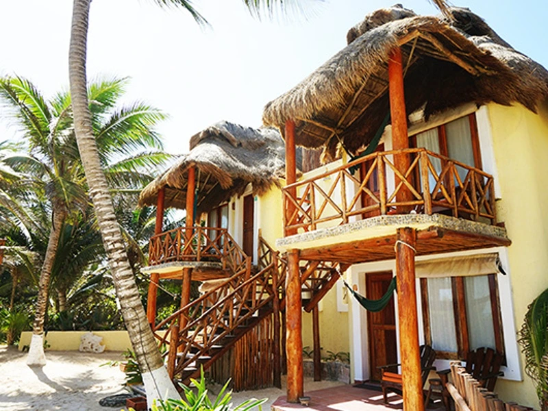 Een strandhuis met rieten dak en houten balkons, omringd door palmbomen, met stoelen en een hangmat voor de ingang.