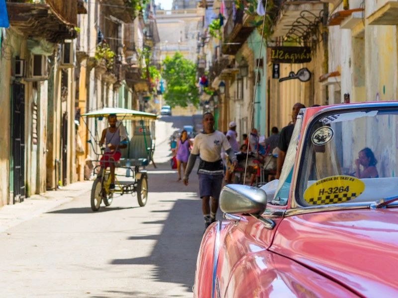 Een levendige straat in een oude wijk met mensen die lopen en fietsen, een fietstaxi en op de voorgrond een klassieke roze auto.