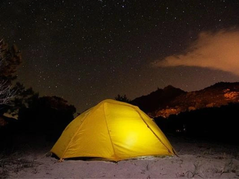 Gele tent die van binnen verlicht is, opgezet in een bergachtig landschap onder een heldere sterrenhemel.