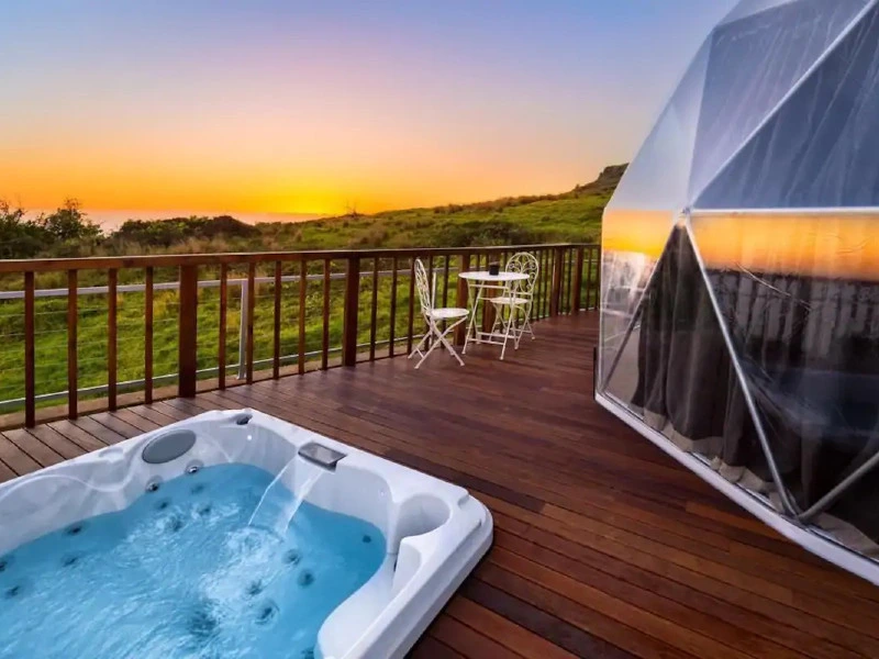 Luxe glampingaccommodatie met houten terras, een jacuzzi en een ronde koepeltent, uitkijkend over groene heuvels bij zonsondergang.