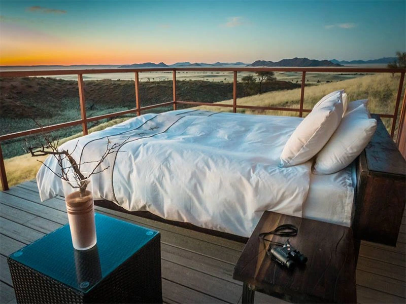 Buitenbed met witte lakens op een houten terras met uitzicht op grasvlaktes en bergen bij zonsondergang.
