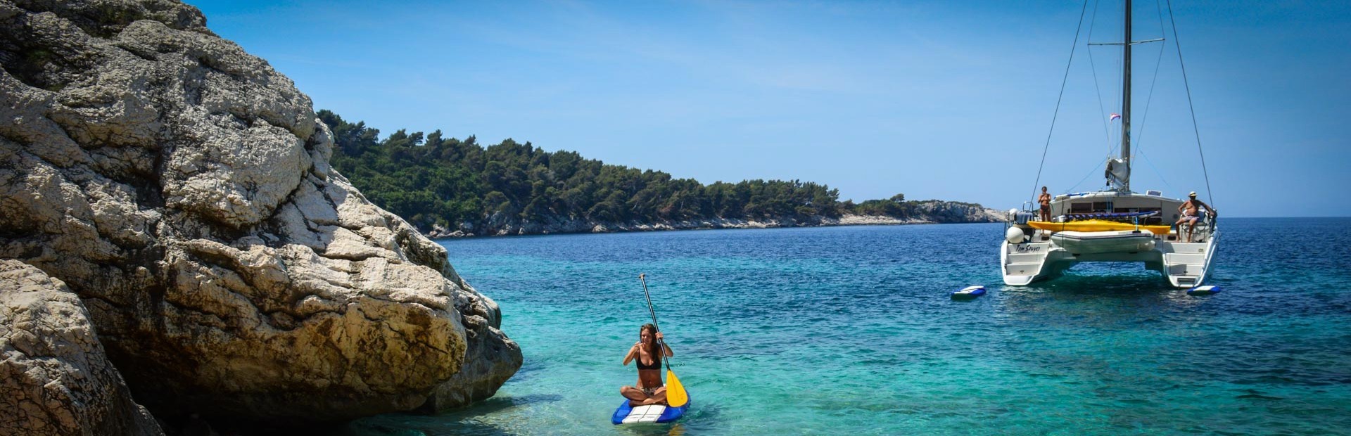 Vrouw peddelt op een SUP-board bij een rotsige kust met een catamaran op de achtergrond in helderblauw water.