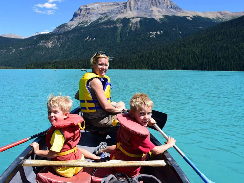 Twee jonge jongens en een vrouw kanoën op een turquoise meer, met hoge bergen en een dicht bos op de achtergrond onder een blauwe lucht.